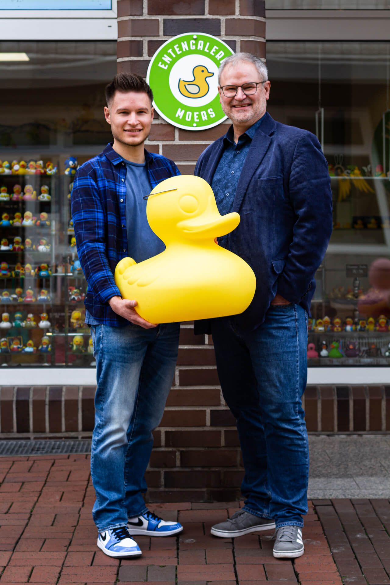 Die Inhaber der Entengalerie Moers stehen mit einer gelben XXL Ente vor dem Schaufenster ihres Badeenten Laden